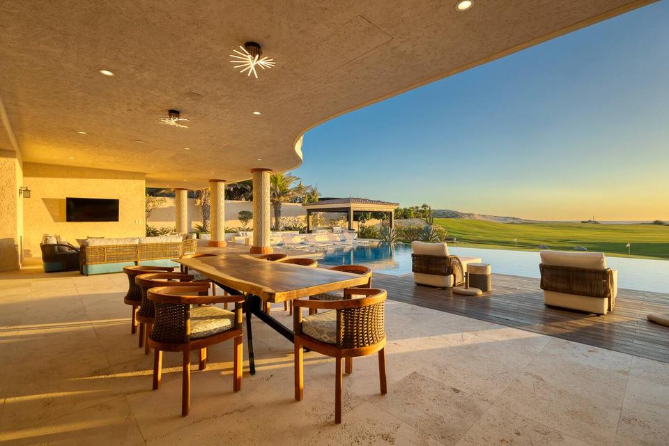 A beautifully designed outdoor living area at the Momentum Cabo property, a Hostaway client, featuring cozy lounge seating, an infinity pool, and scenic golf course views bathed in the warm glow of a stunning sunset.