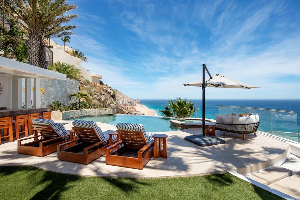 A stunning view of the Momentum Cabo property, a proud Hostaway client, highlighting a serene infinity pool, elegant sun loungers, a stylish outdoor seating area, and breathtaking ocean views against a vibrant blue sky.