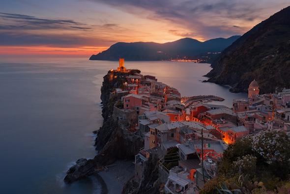 A dramatic dusk view of Vernazza, one of the five villages of Cinque Terre, perched on coastal cliffs with warm golden and amber lights illuminating the historic buildings against a vivid sunset sky — capturing the kind of high-demand Italian destination where local short-term rental restrictions and compliance requirements are most actively enforced.
