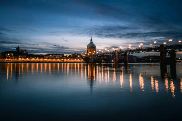 The Pont Saint-Pierre glows softly at dusk in Toulouse, its lights reflecting beautifully on the serene Garonne River, creating a captivating evening view.