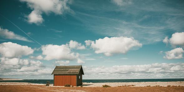 A lone red beach hut sits on a deserted shoreline with no visitors in sight—a visual metaphor for the quiet periods that coastal vacation rental properties experience during the off-season. The vast, empty landscape and open sky emphasize how dramatically demand can drop when peak travel months end, leaving properties waiting for their next booking.