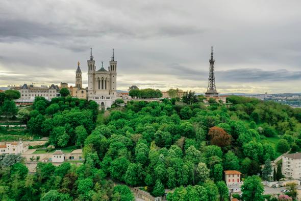 An image of the Basilica of Notre-Dame de Fourvière and the Metallic Tower of Fourvière rising above lush green hills under a cloudy sky in Lyon, France.