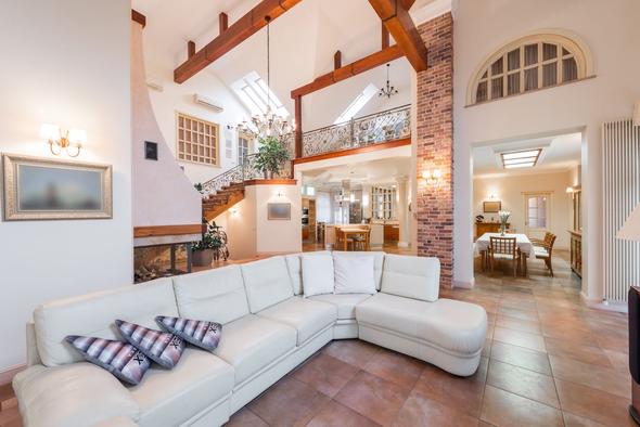 This image captures a grand living space with towering arched windows framed in warm wood, flooding the room with natural sunlight. A crystal chandelier hangs from the double-height ceiling above white sofas and a glass coffee table on polished marble flooring. Through the windows, a manicured garden with mature trees and a brick patio is visible. This image exemplifies the kind of professional, high-resolution property photography that act as a direct ranking signal on Vrbo — properties with compelling photo libraries outperform competitors regardless of other listing factors