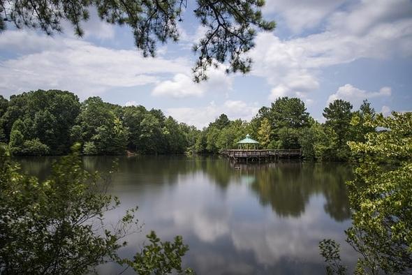 Peaceful lake scene in Roswell, Georgia, with a wooden gazebo overlooking the water, surrounded by lush greenery and a partly cloudy sky.