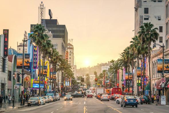 A bustling view of Hollywood Boulevard in Los Angeles, featuring bright neon signs, palm trees lining the streets, and a golden sunset casting a warm glow over the busy urban landscape.