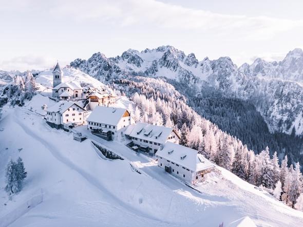 An aerial view of a snow-dusted mountain village in the Italian Alps, surrounded by frost-covered pine trees and dramatic rocky peaks — representing Italy's northern alpine regions where short-term rental rules and tourist tax rates may differ significantly from urban centers.